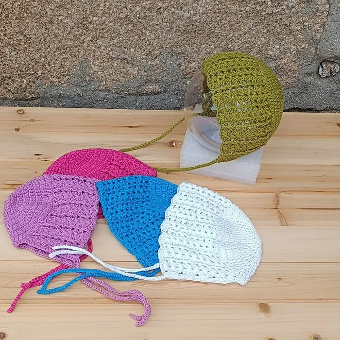 Capota clásica "Sirope" para bebé en crochet con cordones. Se muestran también varias unidades equivalentes en tonos fucsia, lila, azul y blanco.